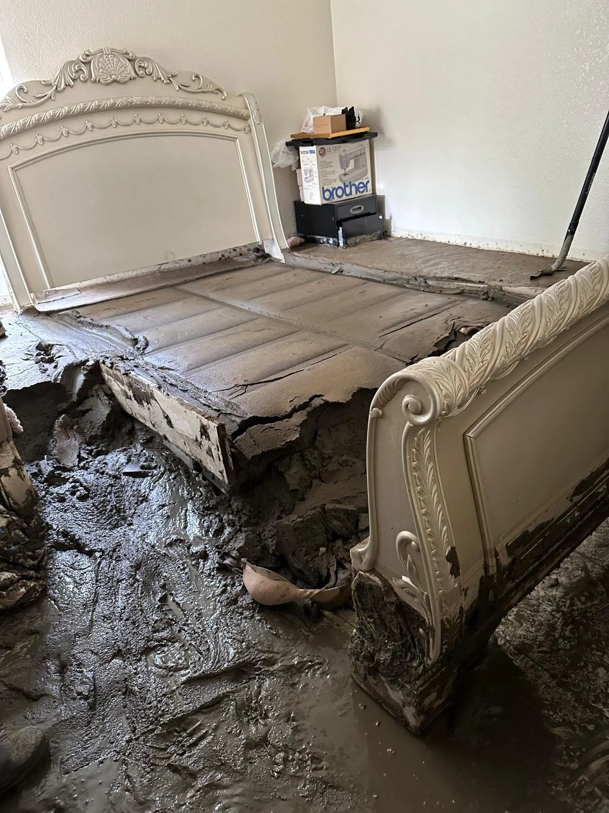 A bedroom with ornate white bed frame removed, showing damaged floor foundation and mud and debris on the ground, indicating a house flood or foundation problem. A box labeled 'Brother' and various items are on the side.