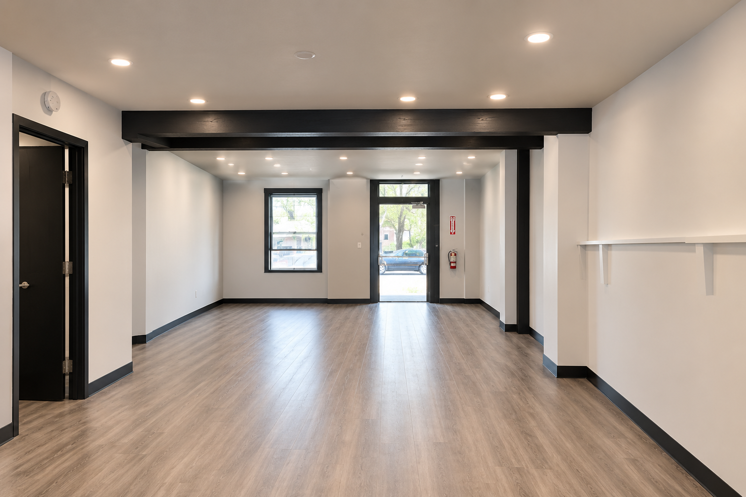 Empty room with wood flooring, white walls, black window and door frames, black beam ceiling, and ceiling lights, with a fire extinguisher on the wall near the door.