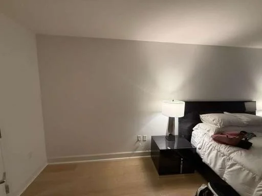 A cozy bedroom with a sloped ceiling, a black nightstand, a white lamp, a bed with white linens and pillows, and a folded piece of clothing on the bed.