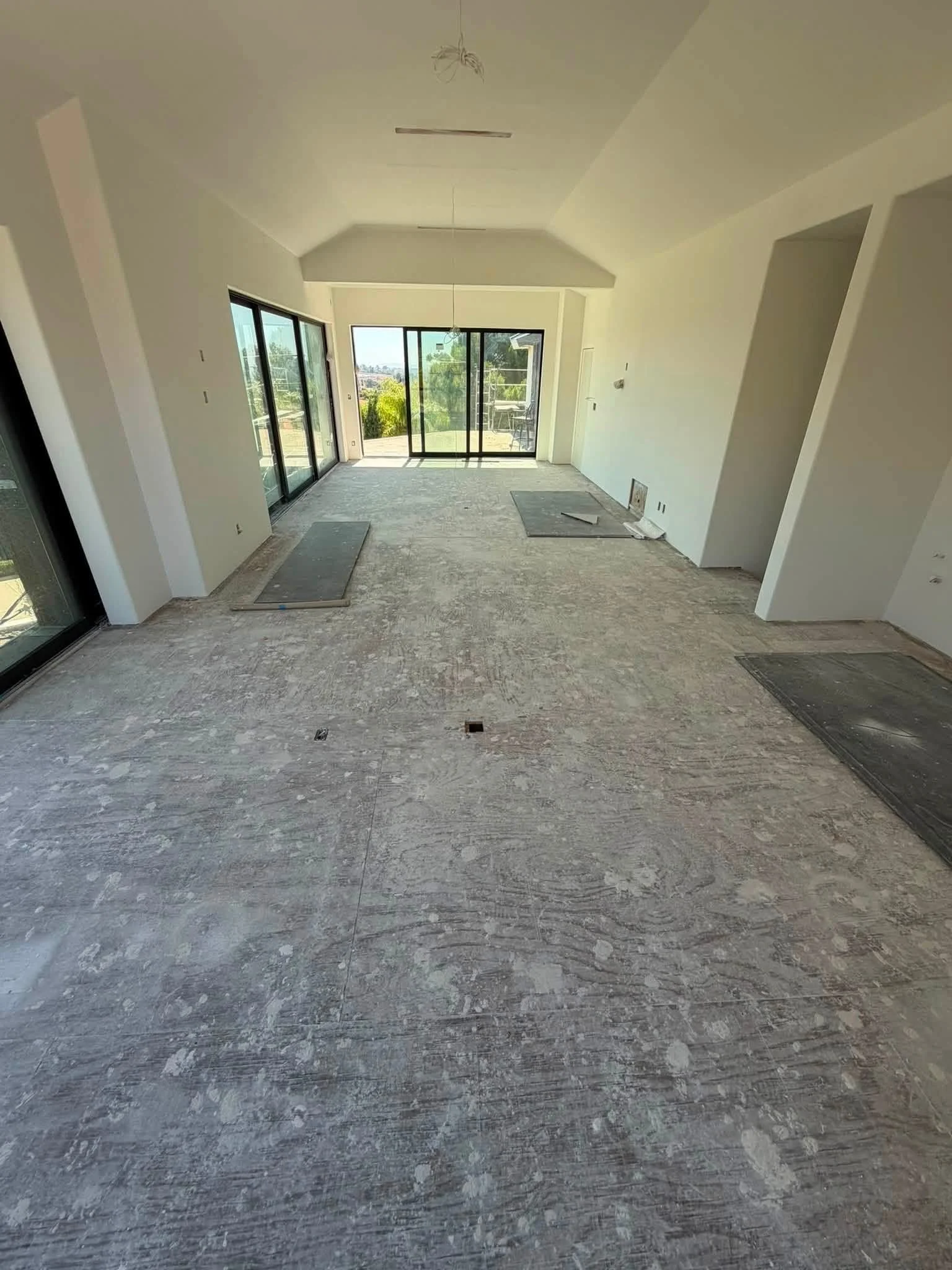 An unfinished room with sliding glass doors leading to an outdoor balcony. The floor is made of gray, textured material. There are some construction materials and tools visible on the floor, and the walls and ceiling are painted white.