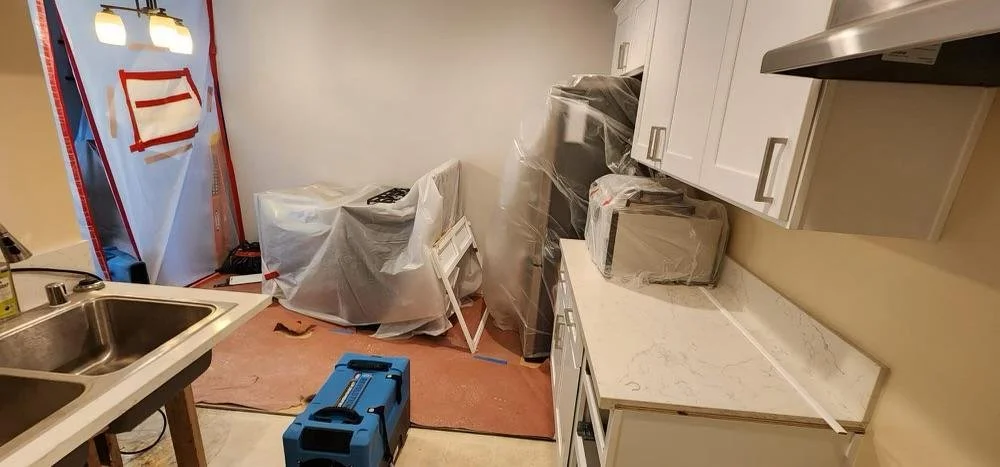 Kitchen under renovation with cabinets, countertop, and covered appliances, plastic sheeting, and a toolbox on the floor.