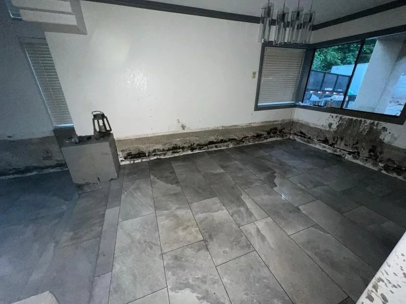 Empty room with damaged walls and mold around the base of the walls, large window with a view outside, gray tiled floor, and a small bottle on a ledge.