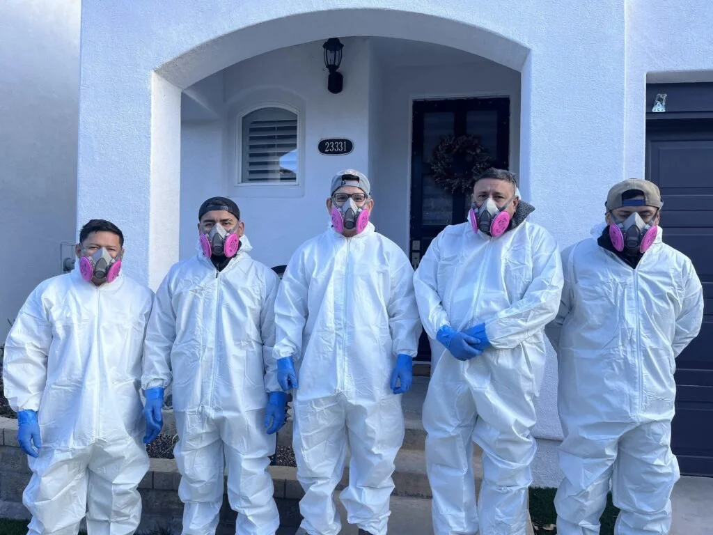 Five people wearing white protective suits, face masks with pink filters, and blue gloves standing in front of a house with the number 23331 on it.