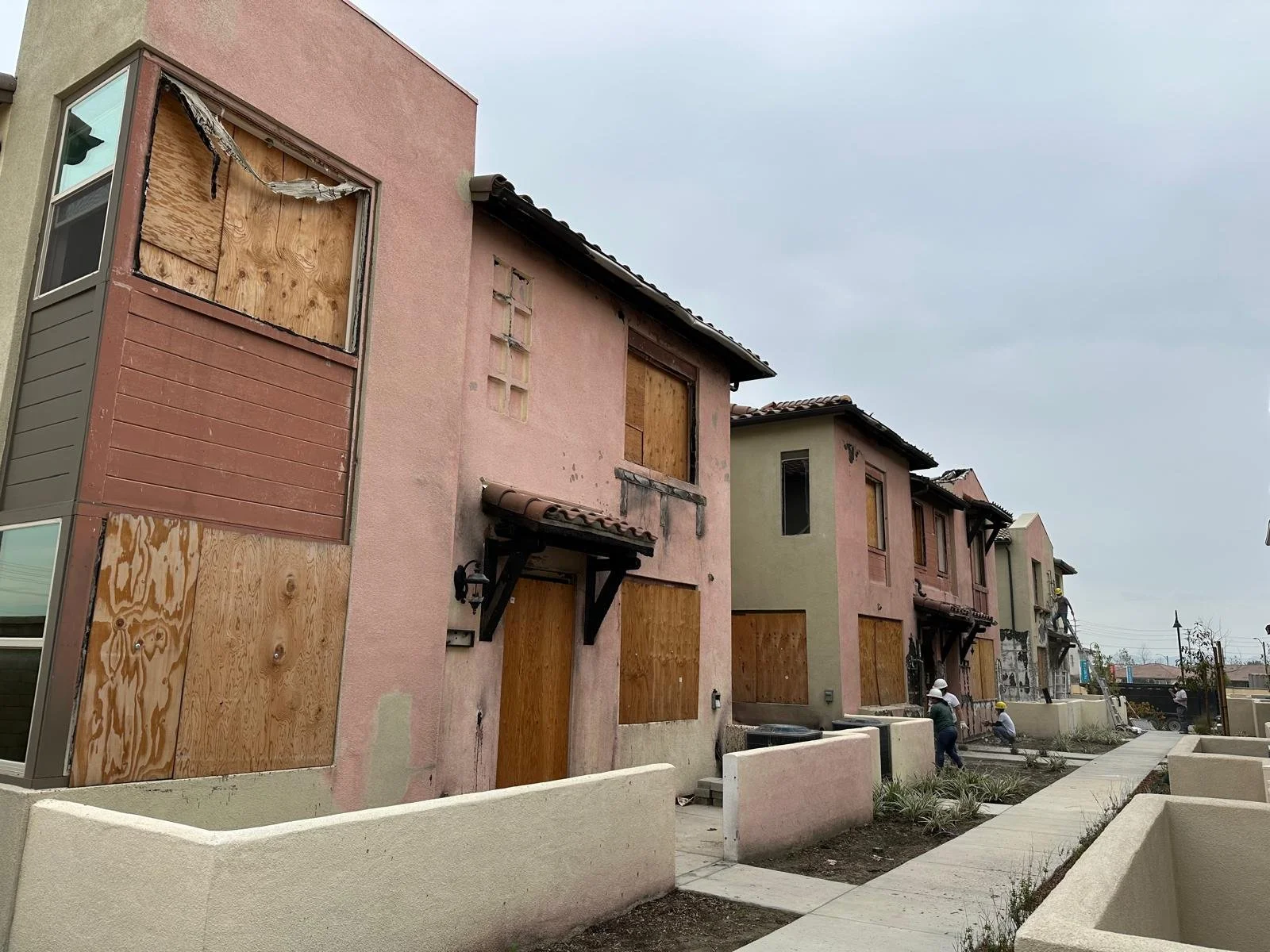 Row of residential buildings with boarded-up windows, construction workers working on the exterior walls, and a sidewalk in front.