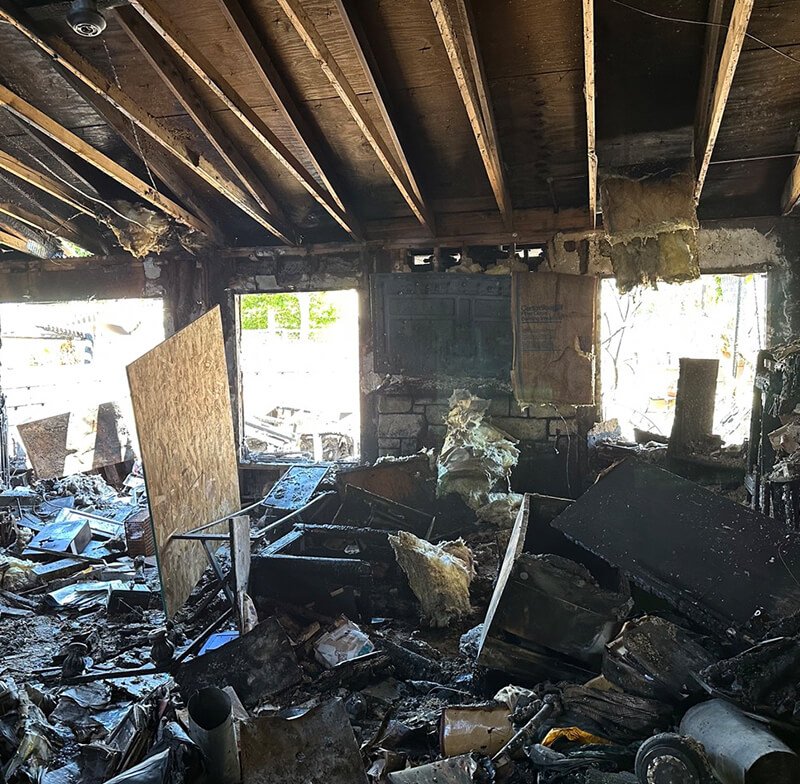 Interior of a burned building with charred debris, damaged furniture, and burnt insulation, with sunlight coming through broken windows.