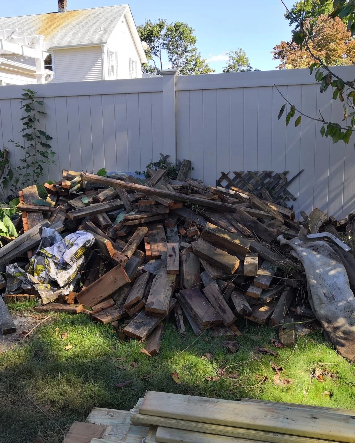 A backyard with a pile of split wooden logs and some lumber, bordered by a white fence, with a house and trees in the background.