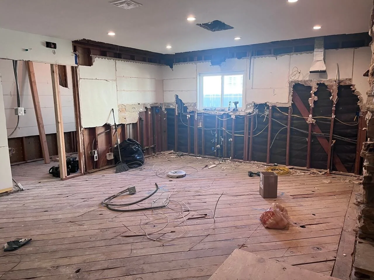 Interior of a room under renovation with exposed wall studs, partially removed drywall, a window, and construction tools spread on the wooden floor.