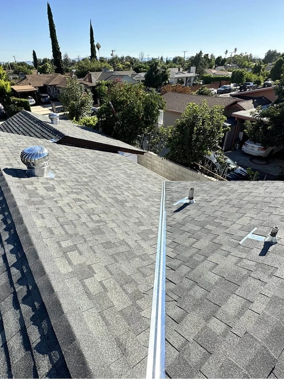 View from the roof of a residential house showing shingles, roof vents, a metal seam, trees, neighboring houses, and a clear blue sky.