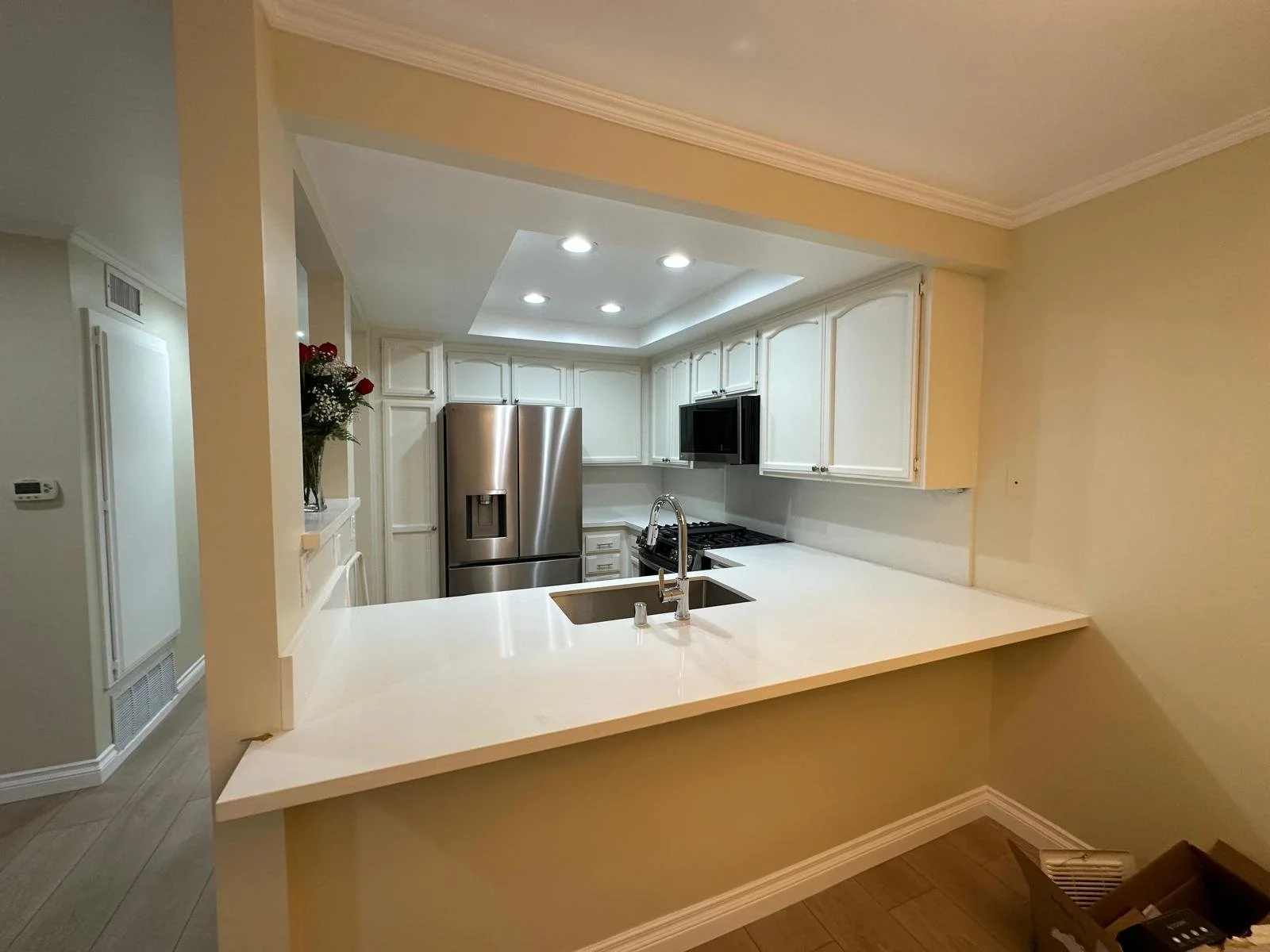 Modern kitchen with white cabinets, stainless steel refrigerator, black microwave, gas stove, white countertops, and a double sink, illuminated by ceiling lights.