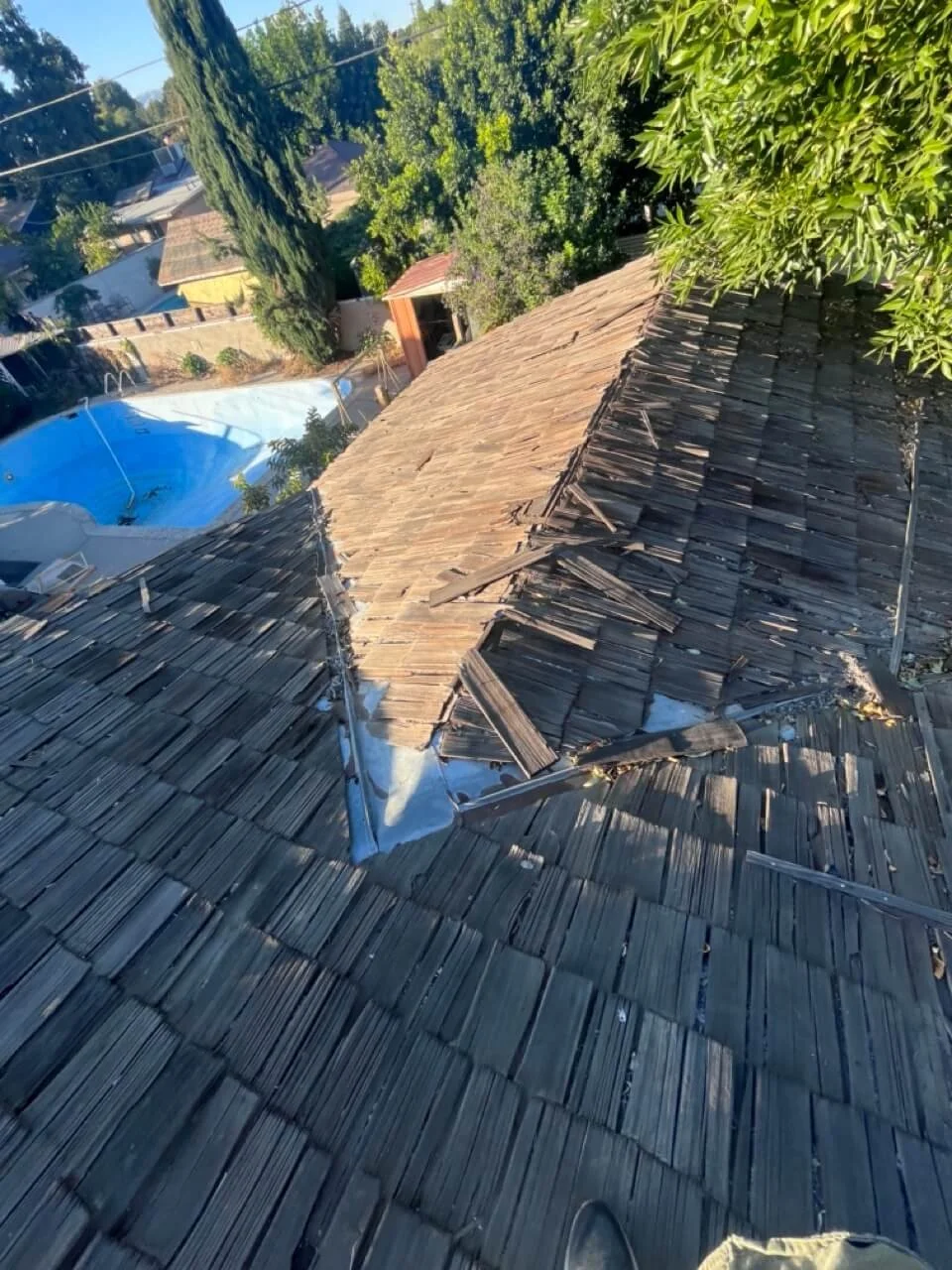 View from a rooftop showing damaged wooden shingles with a section of the roof torn off, revealing the underlying structure, along with surrounding trees and neighboring houses.
