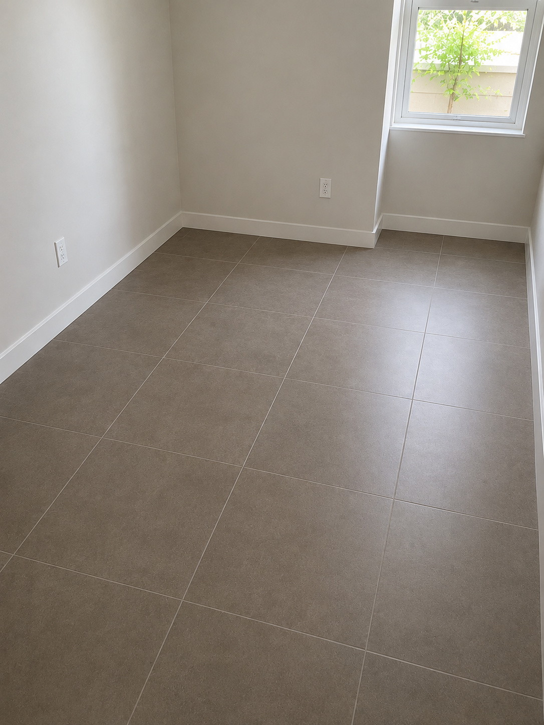 Empty room with beige tiled flooring, white walls, a window showing greenery outside, and electrical outlets on the wall.