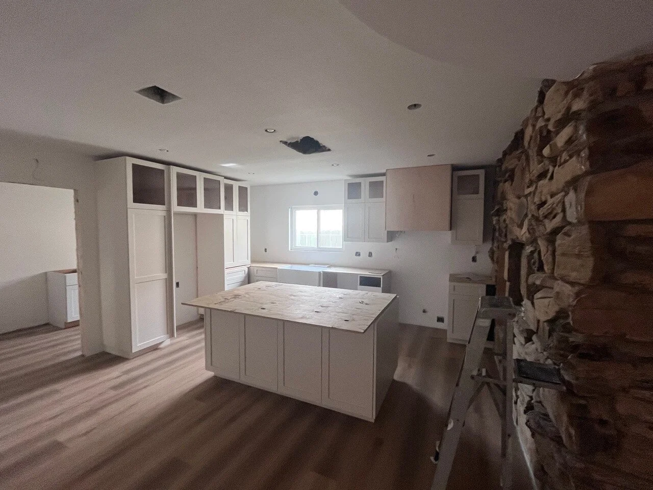 Kitchen under renovation with unfinished cabinets, an island, and a stone feature wall, with a ladder and construction materials.