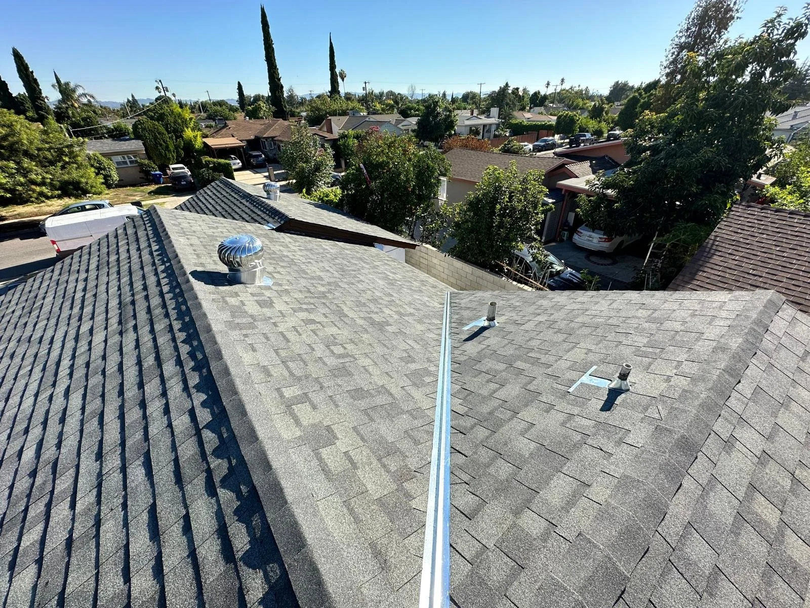A view from the rooftop showing asphalt shingles, roof vents, and neighboring houses with trees and clear blue sky in the background.