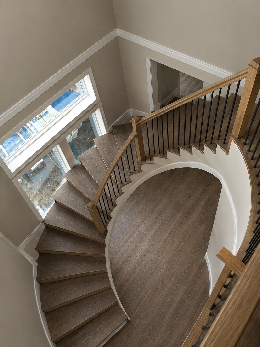 View of a curved wooden staircase with black metal balusters and a wooden handrail, leading downstairs, next to a wall with large windows allowing natural light into the space.