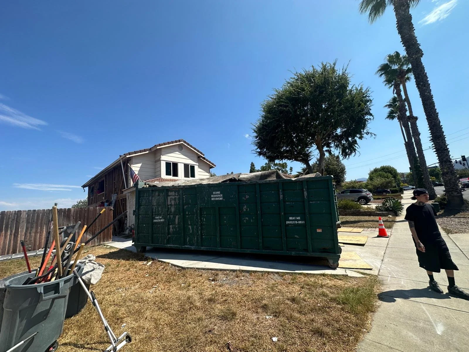 Construction site in a suburban area with a partially renovated house, large green dumpster, construction tools, and a woman walking along the sidewalk, with palm trees and parked cars in the background.