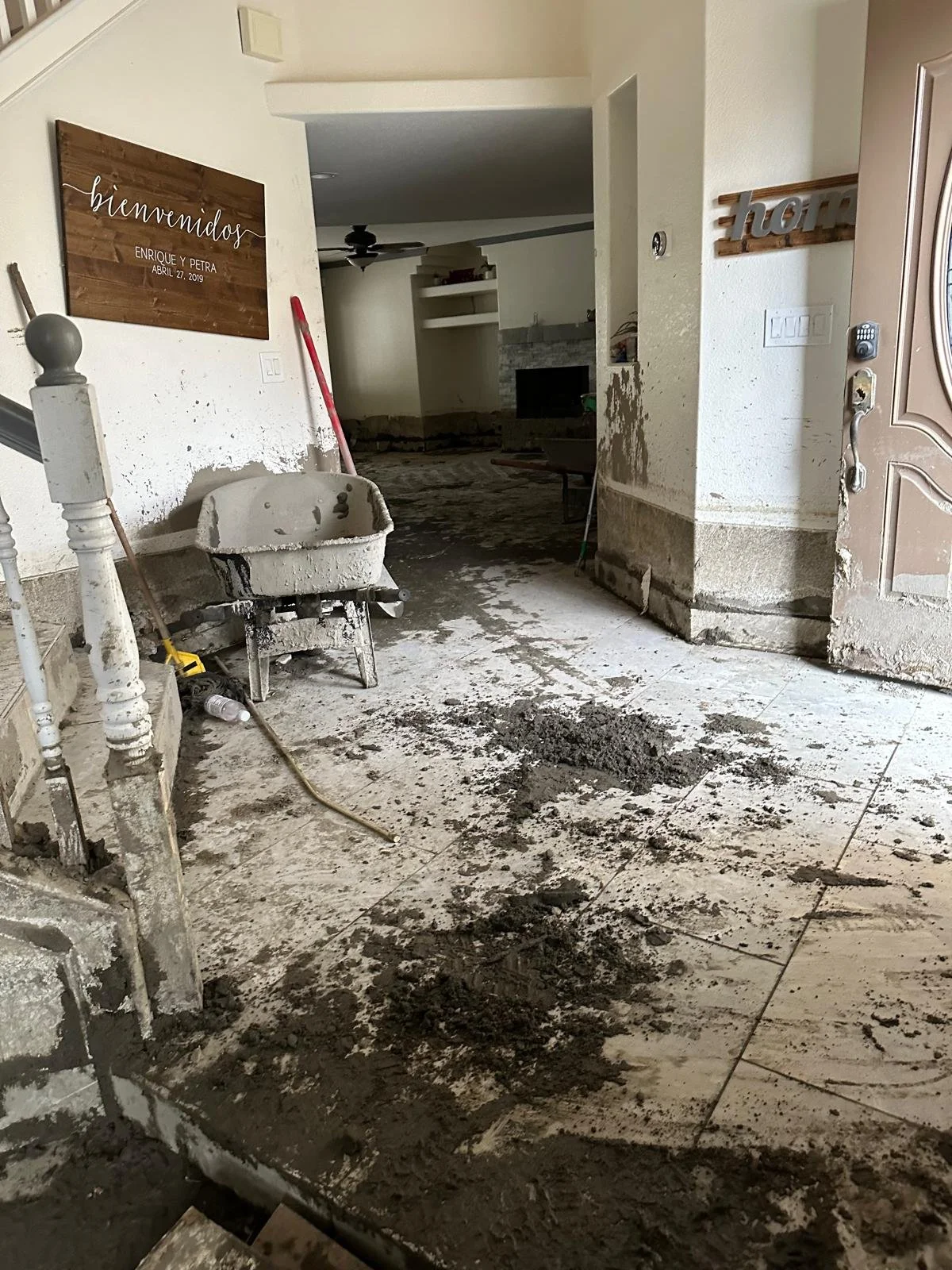 Interior of a house under renovation with construction debris, a wheelbarrow, and tools on the floor near a staircase, with walls partially damaged and a sign that reads 'bienvenidos' on the wall.