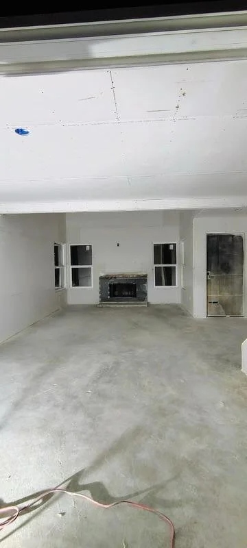 Empty room under construction with white walls, a fireplace, and three windows, unfinished concrete floor, and some construction wires on the floor.