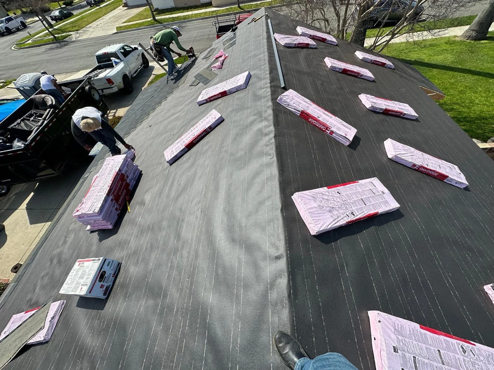 Workers installing roofing shingles on a house roof. Several bundles of shingles are spread across the roof, and a truck and a trailer filled with materials are parked nearby. The scene is in a residential neighborhood.