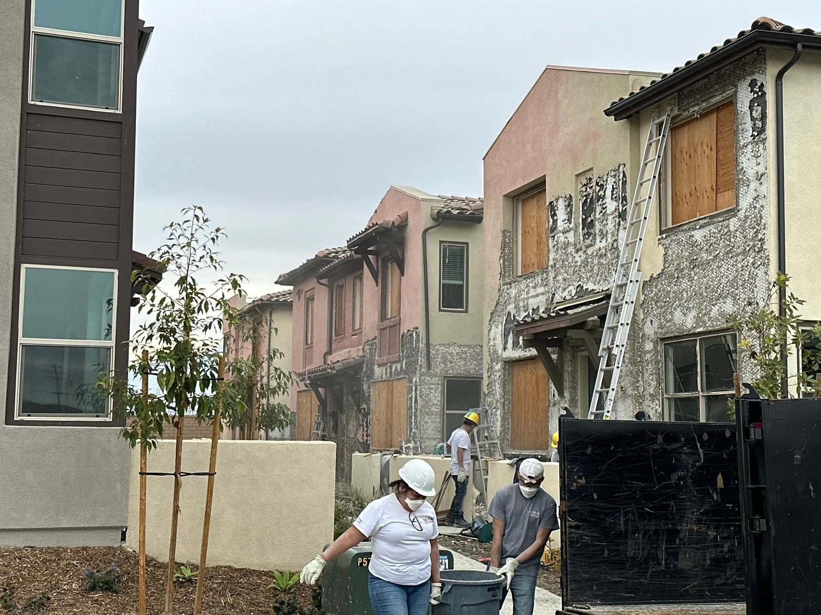 Construction workers and ladders repair damaged building facades with boarded-up windows in a residential area on a cloudy day.