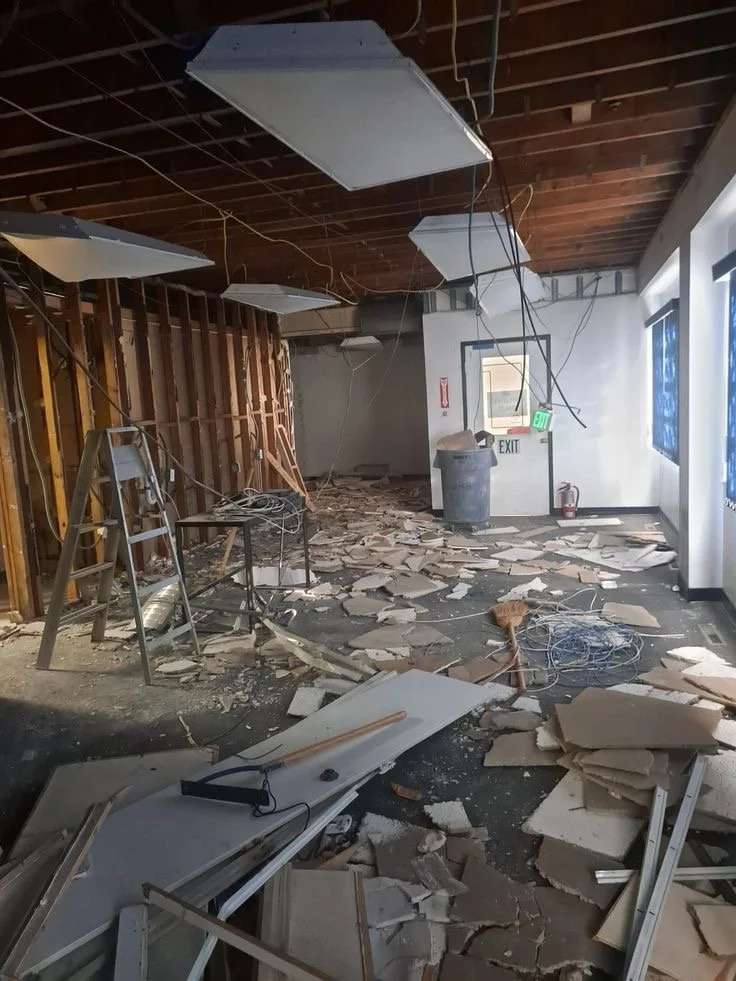 Interior of a building with ceiling tiles on the floor and hanging from the ceiling, exposed wires, a ladder, and debris scattered across the floor, indicating extensive damage or construction.