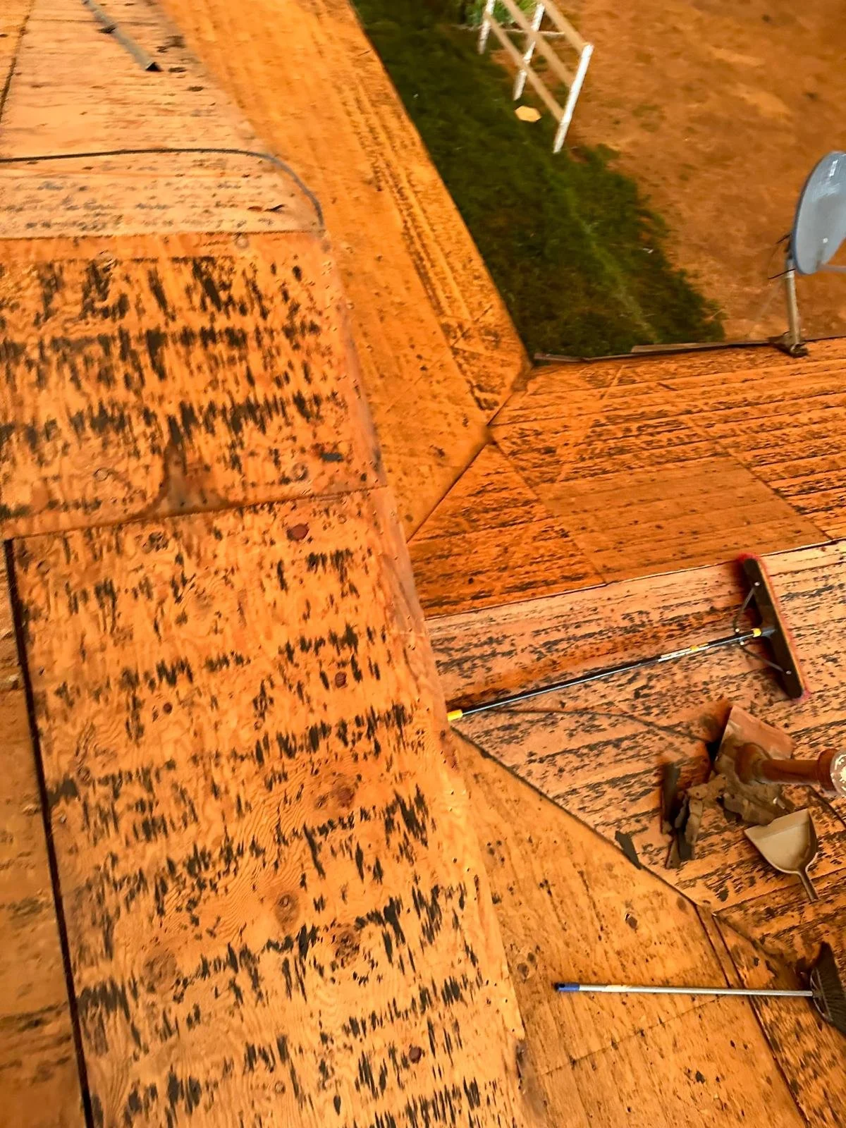 Close-up of a wooden deck with some tools, including a squeegee and a scraper. The deck appears to be in the process of being cleaned or sanded, with a ladder and some grass visible in the background.