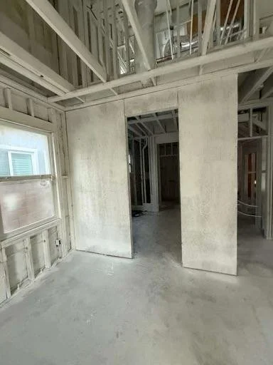 Interior of a house under construction with exposed framing, drywall, and unfinished walls. A doorway leads into another room, and a window is visible on the left side.
