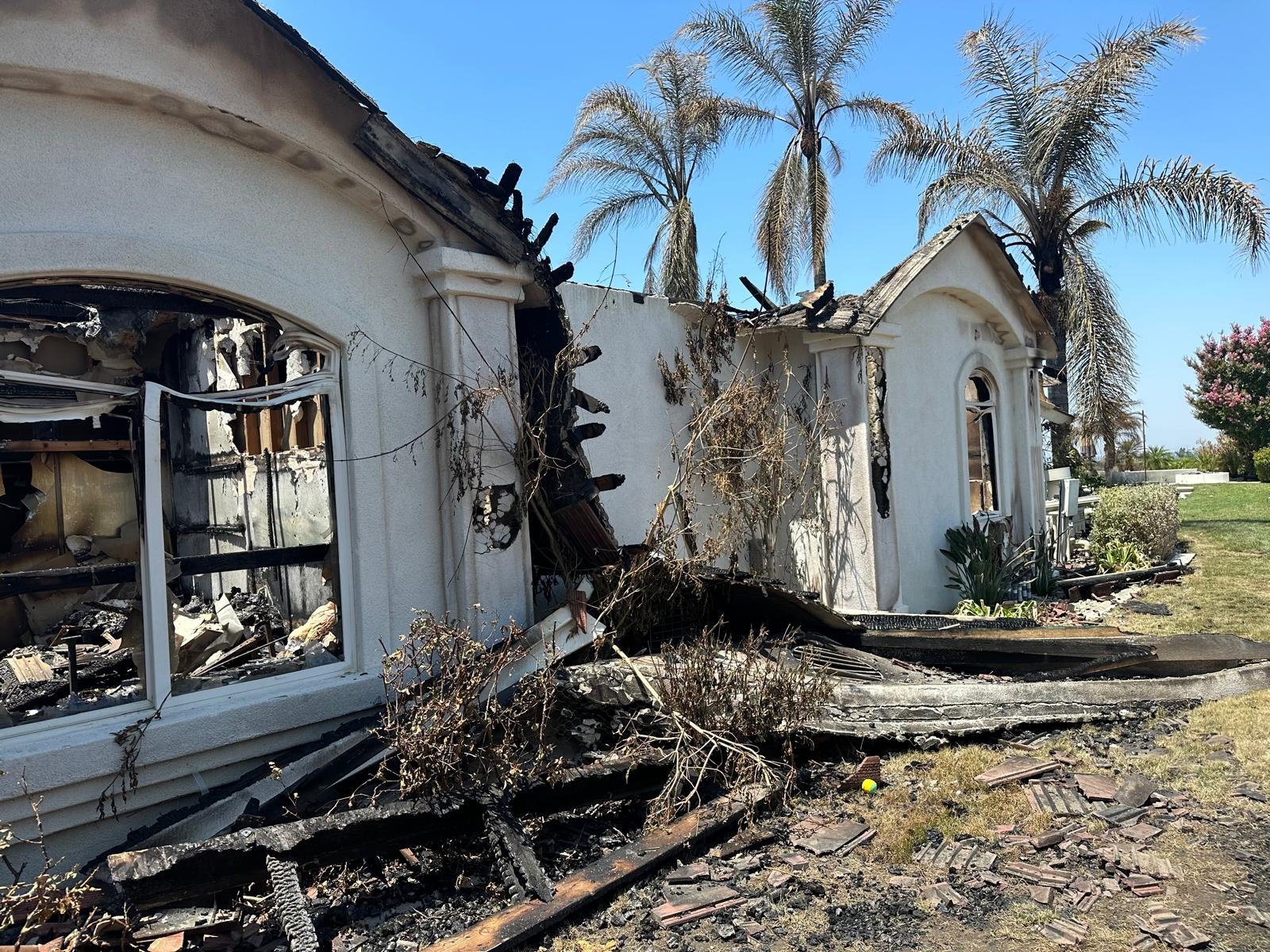 A house damaged by a fire with charred walls and a destroyed roof, smoke damage visible, and debris on the ground