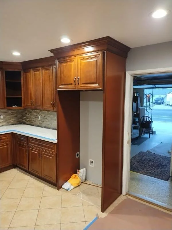 View of a kitchen with wooden cabinets, tiled backsplash, and a countertop, adjacent to a doorway leading to a room with a chair and a rug.