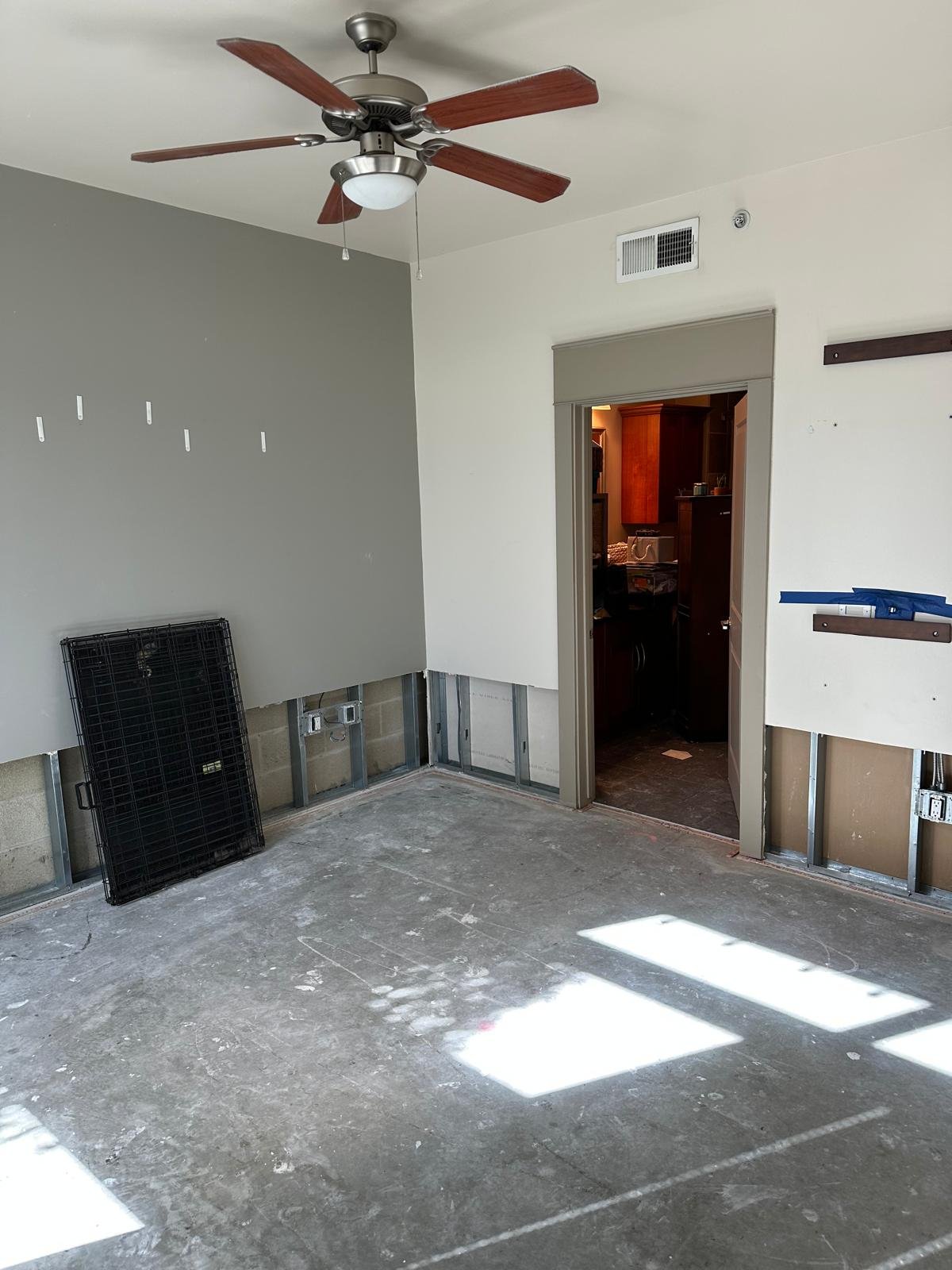 Room undergoing renovation with exposed wall studs, a ceiling fan, and a partially removed wall. The floor is concrete with dust and markings, and there is a black metal grate leaning against the wall.