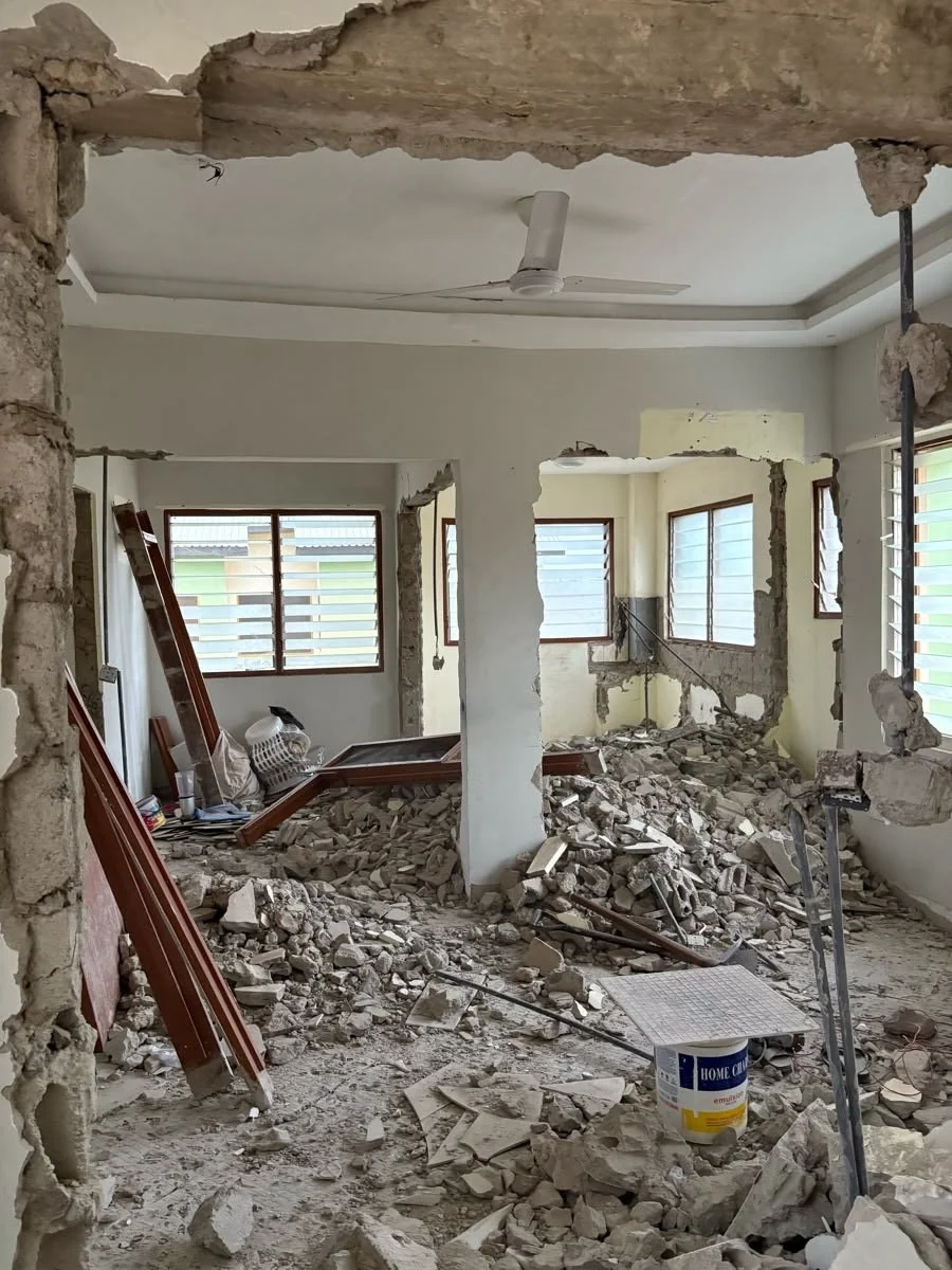 Interior of a house undergoing renovation with rubble and debris on the floor, exposed walls, and construction materials scattered around.