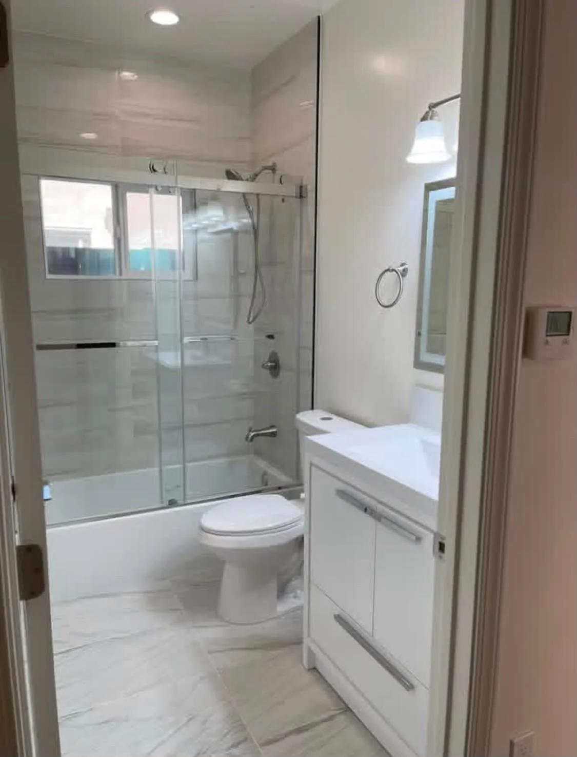 A small modern bathroom with a glass enclosed shower, white toilet, small white vanity with a sink, mirror, and a window, with beige tile flooring and walls.
