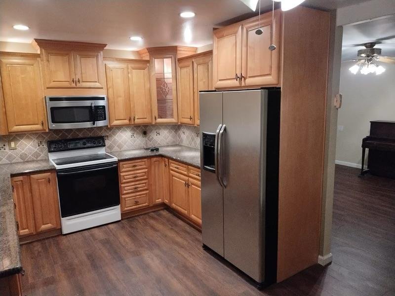 Kitchen with wooden cabinets, stainless steel refrigerator, white stove with black trim, microwave, and tiled backsplash. Dark hardwood floors and a partial view of another room with a piano.