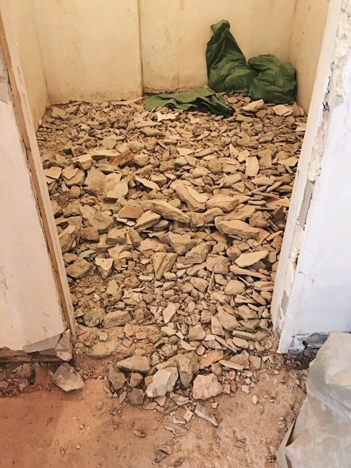 Room under renovation with drywall debris and rocks on the floor, and green bags in the corner.