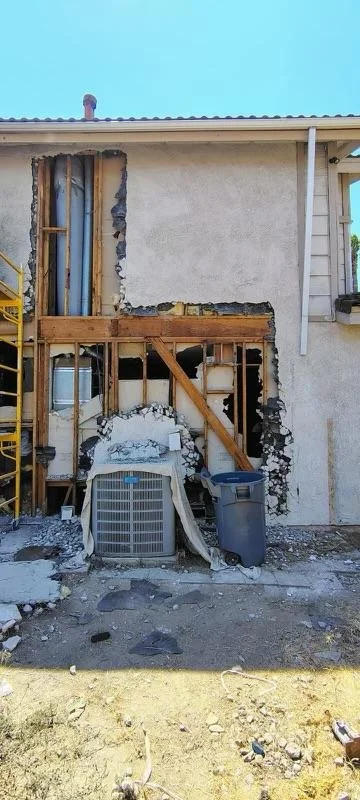 Partially demolished exterior wall of a house revealing framing, plumbing, and an air conditioning unit, with construction debris on the ground.