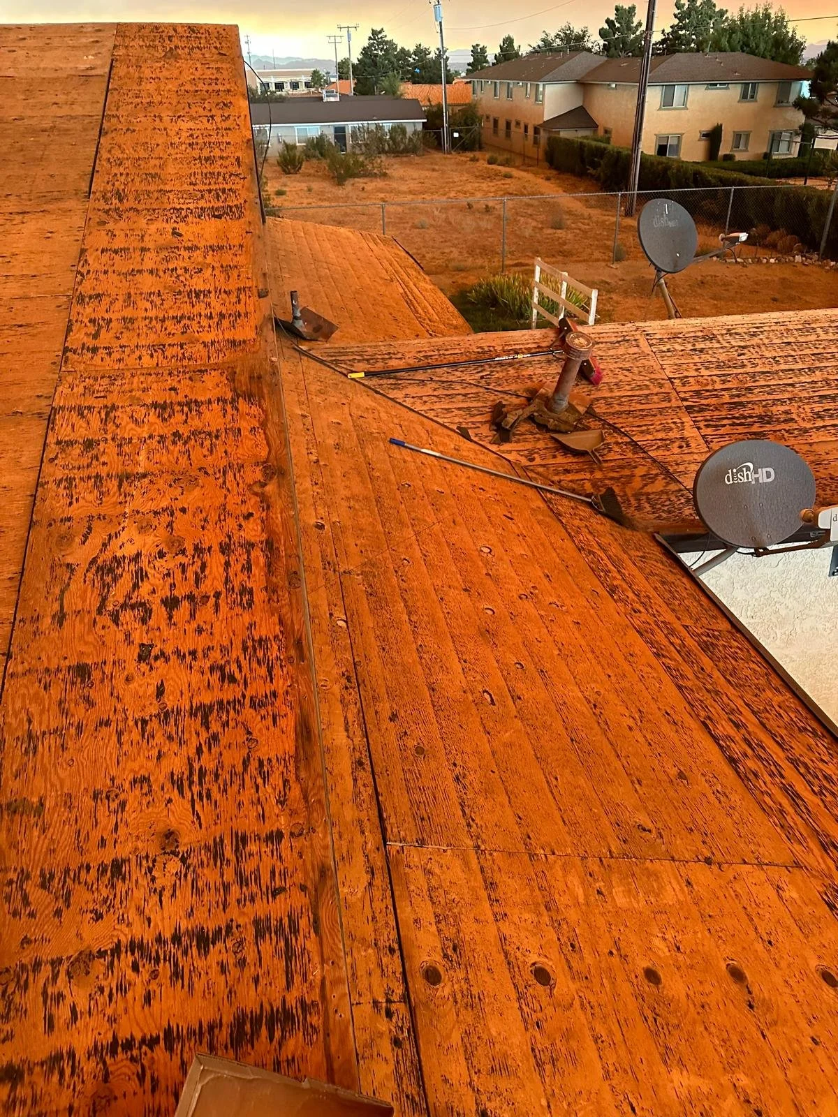 View of a wooden rooftop under construction with satellite dishes, colored in a warm orange hue, overlooking a residential neighborhood with houses, fences, trees, and utility poles.