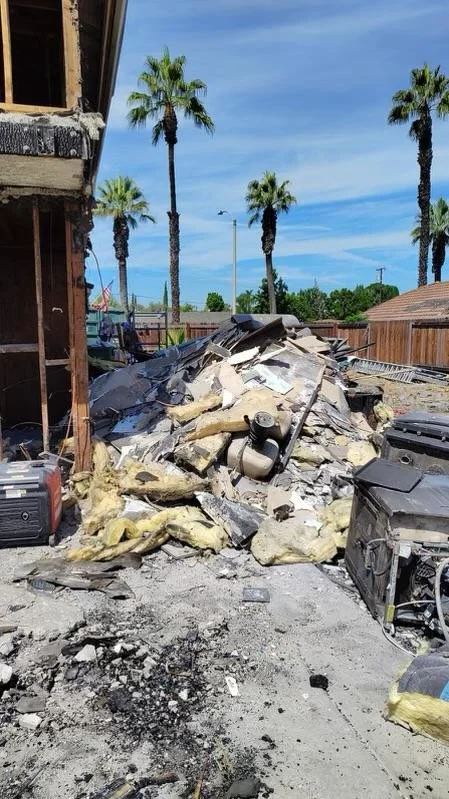 Exterior of a house damaged by fire with a pile of debris, burnt insulation, and appliances, with palm trees and a clear blue sky in the background.