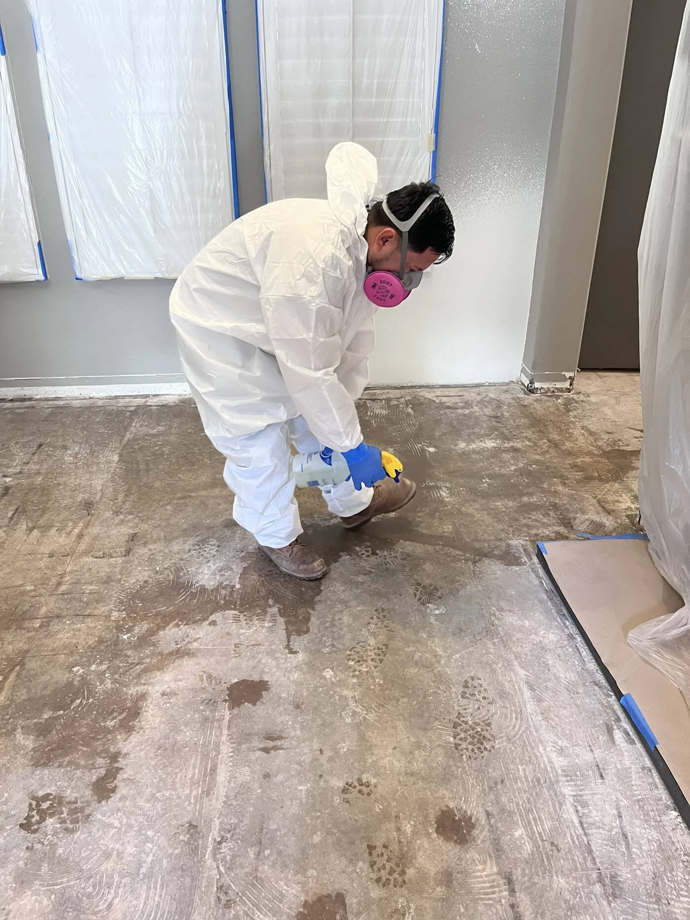 A person wearing a white protective suit, gloves, a pink respirator mask, and goggles is cleaning or preparing a concrete floor in a construction or renovation site. The room has plastic sheeting covering the walls and equipment nearby.