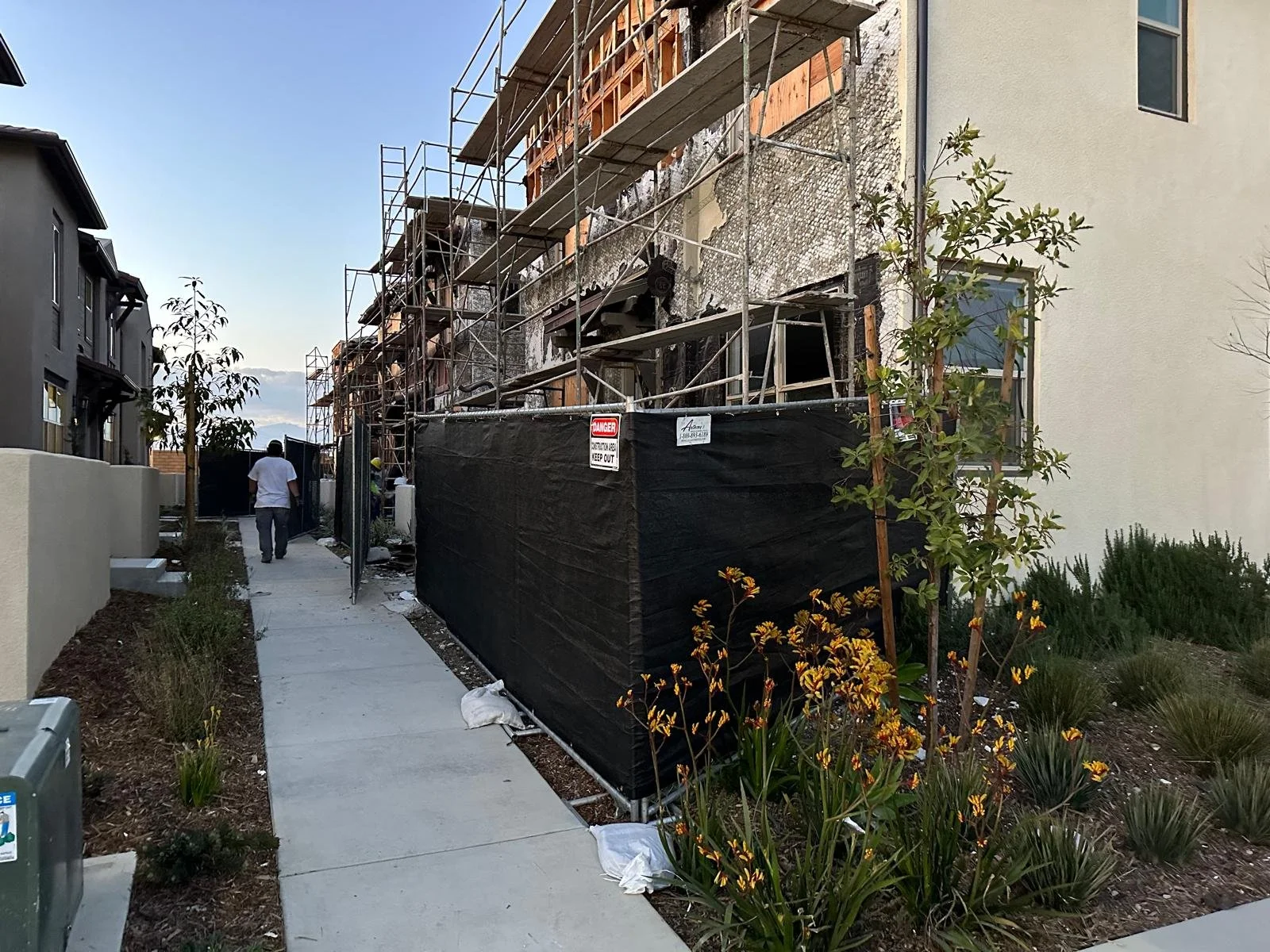 Construction work on a building with scaffolding, partially covered with black fencing, in a residential neighborhood, with workers visible and a pathway lined with plants and flowers.