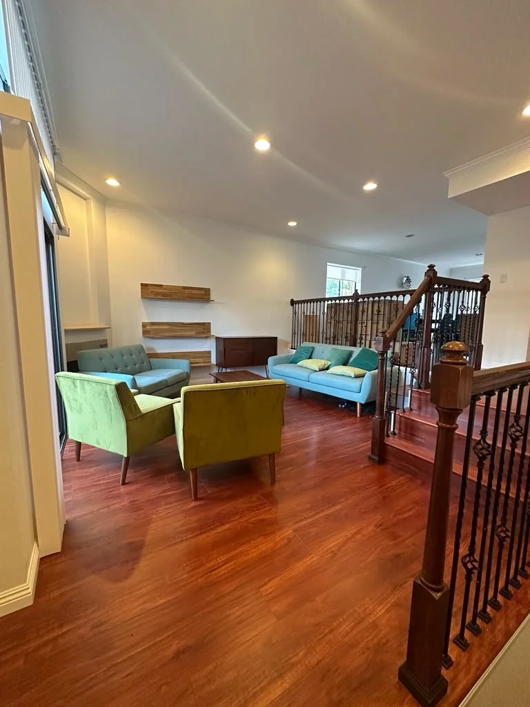 Living room with wooden floors, a wooden staircase with black iron balusters, two light green chairs, two blue sofas, recessed ceiling lights, and wall-mounted wooden shelves.