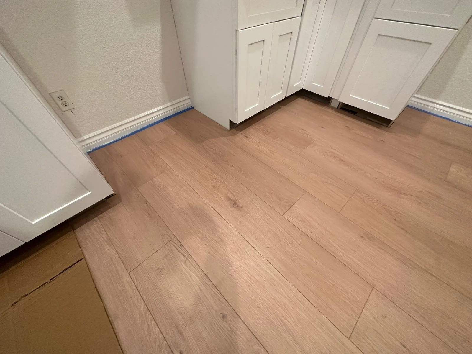 Close-up of a kitchen corner with light wood flooring, white cabinets, beige textured wall, and blue painter's tape along the baseboard.