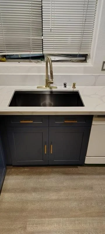 Kitchen sink with a gold faucet and a black basin, set in a white marble countertop. Behind the sink are closed white blinds. There are two cabinet doors painted dark blue with gold handles beneath the countertop.