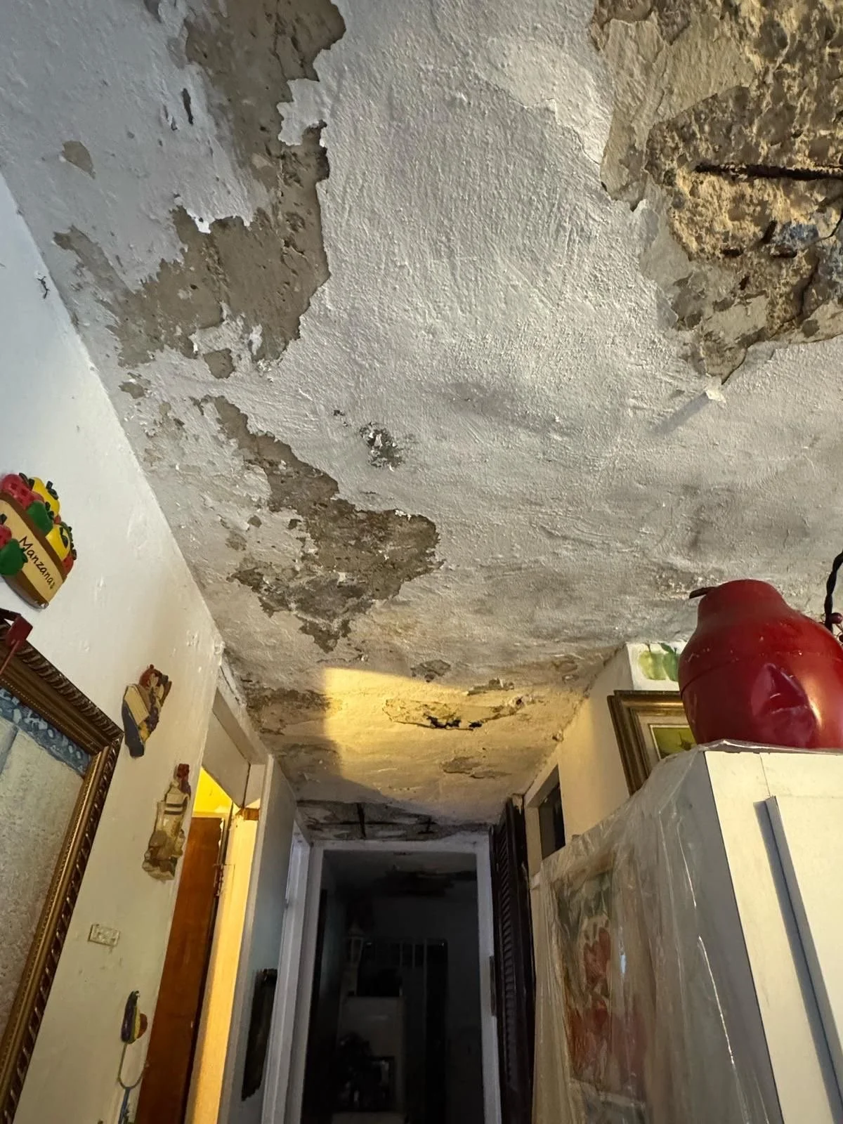 Ceiling with peeling paint and water damage in a dimly lit hallway, with wall decorations and furniture visible.