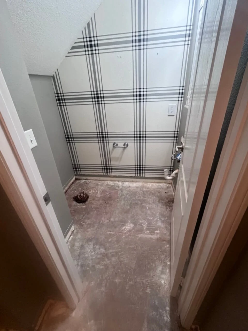 Small room under renovation with unpainted floor, gray painted walls, and graffiti-style black and white plaid wallpaper on one wall. There is a pipe and a grab bar installed on the wall, and a lip at the bottom of the wall indicating a potential sho