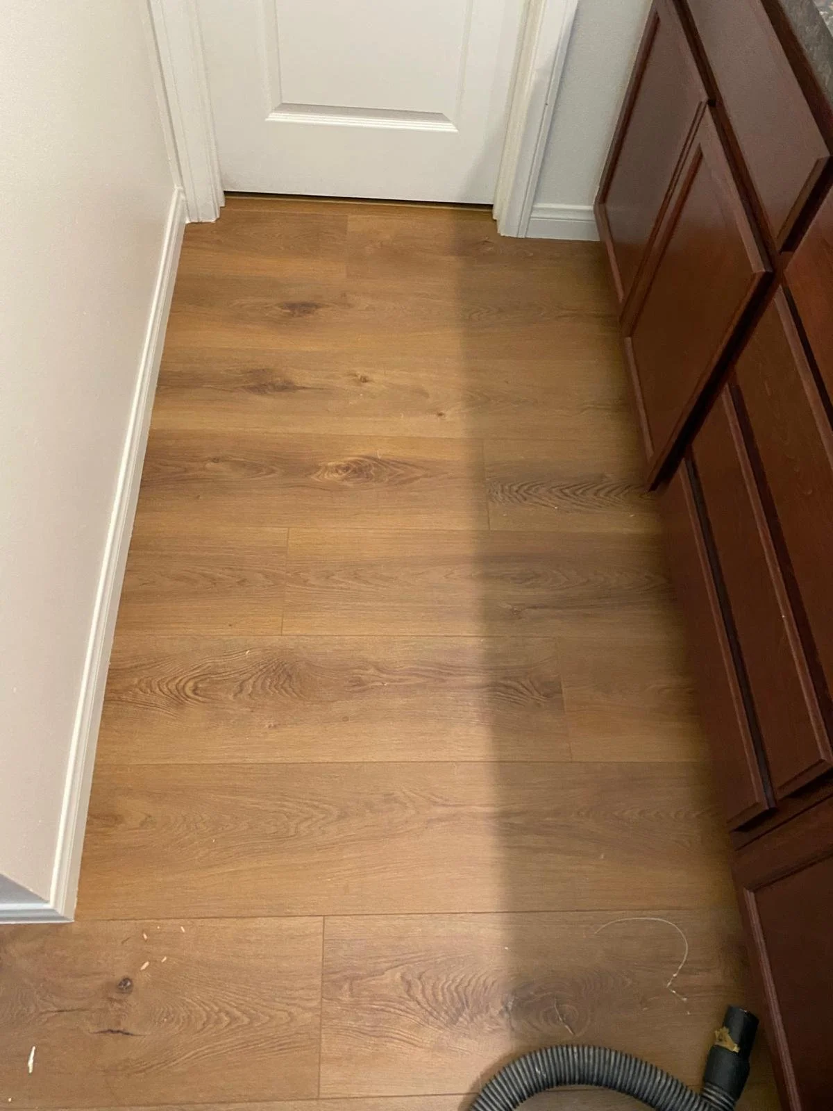 Wooden floor with a white door, white walls, and brown cabinet on the right, with a vacuum hose at the bottom right corner.