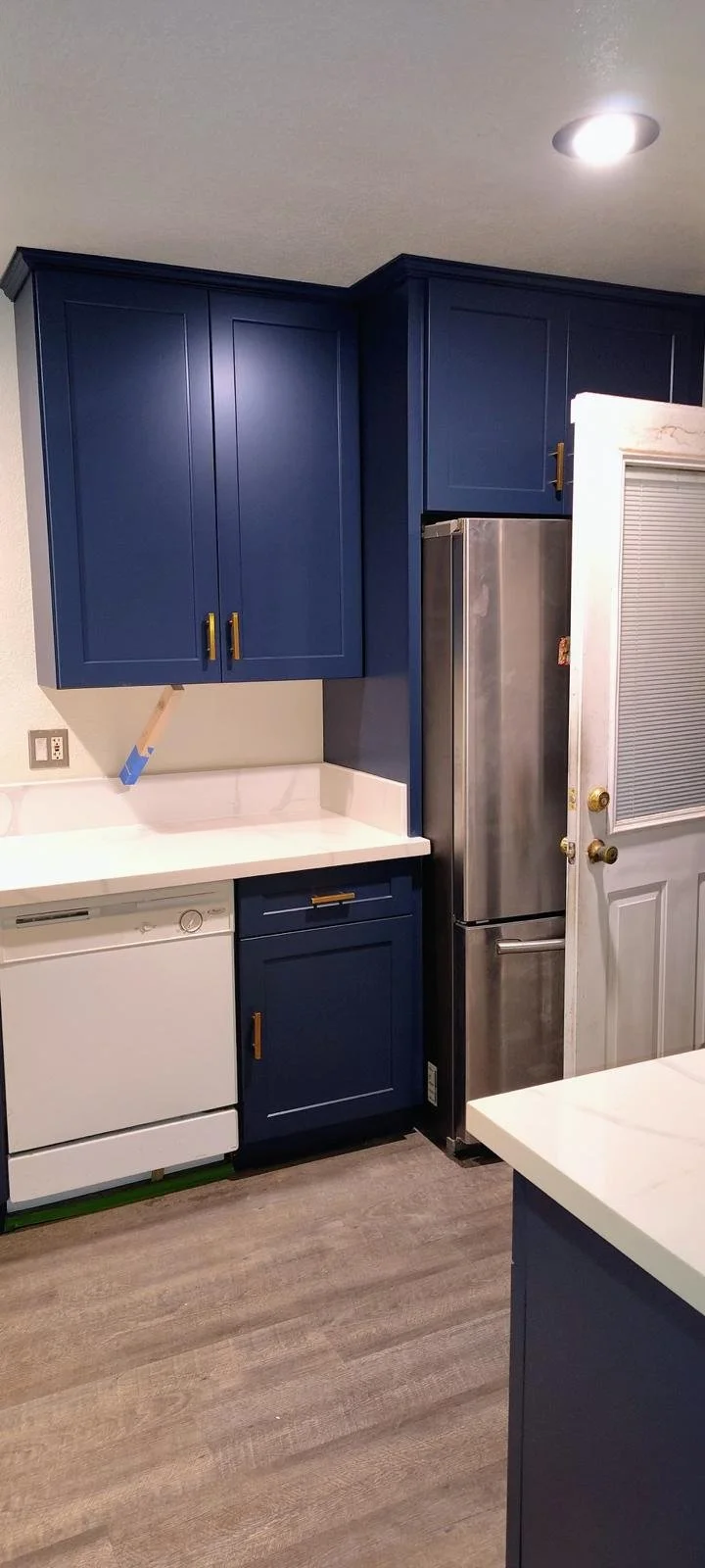 A kitchen with blue cabinets, white countertops, a white dishwasher, a stainless steel refrigerator, and a door with a window blind.