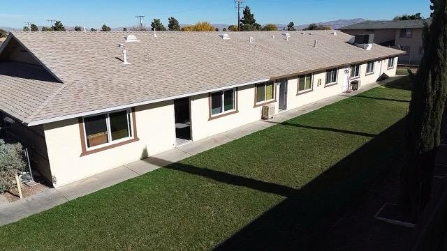 Back view of a single-story apartment or townhouse complex with a beige exterior, brown window frames, and a gray shingled roof. There is a well-maintained lawn in the backyard along with some trees and neighboring buildings in the background.