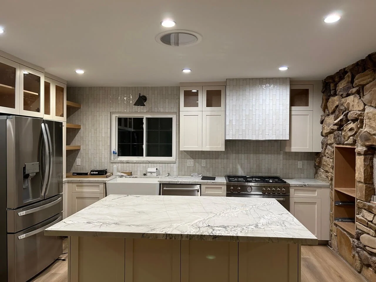Modern kitchen with stainless steel refrigerator, white cabinets, marble island, stone wall, and tiled backsplash.