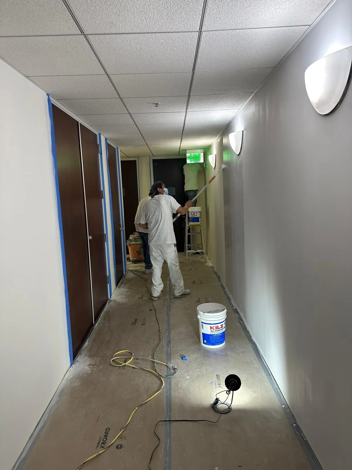 Workers painting a hallway wall, one on a ladder applying paint with a roller, others working nearby, with painting supplies and tape on the floor.