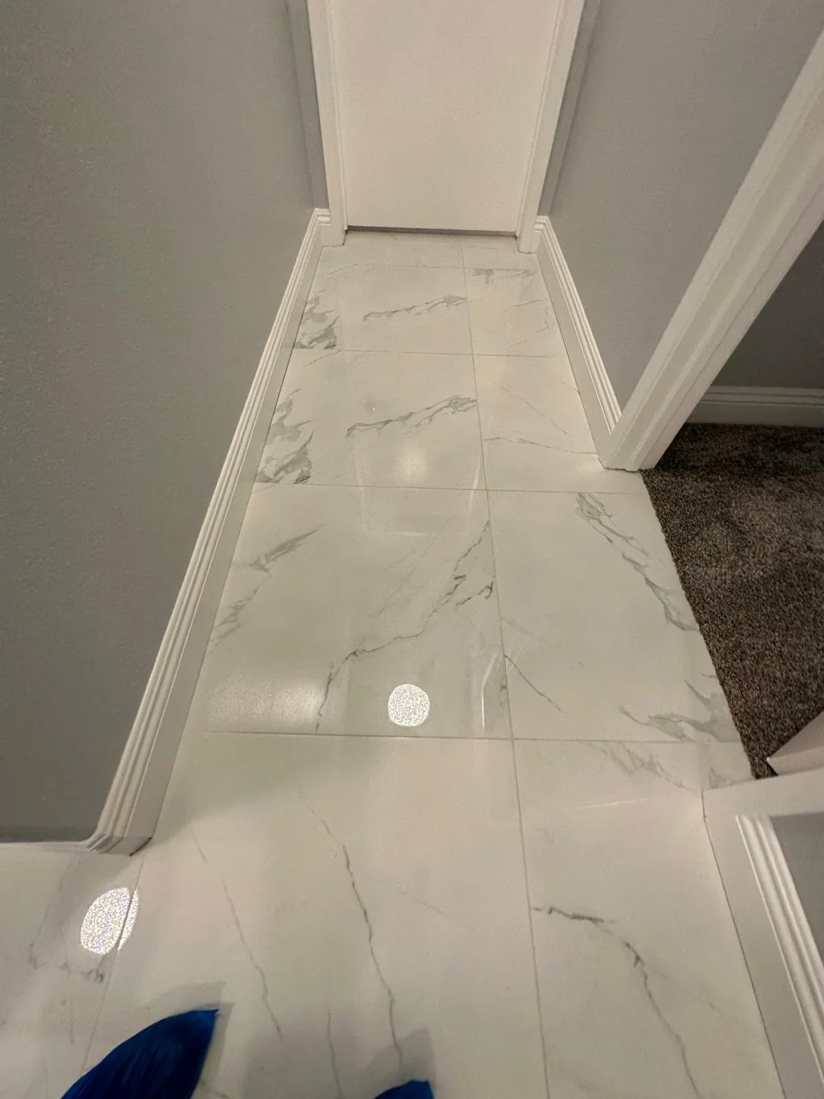 A view of white marble tile flooring with gray veining inside a home, next to a wall with gray paint and white baseboard, and a closed white door.