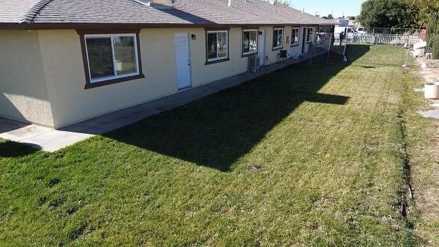Backyard with a grassy lawn, concrete walkway, and a single-story beige house with multiple windows and a door. Shadows cast across the yard from the house and nearby objects.